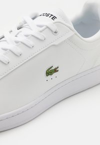 Witte leren sneaker met vetersluiting, voorzien van geborduurd groen krokodil logo en kleine ventilatiegaatjes nabij de zool.