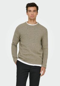 Beige vlněný svetr s kulatým výstřihem a žebrovan�ými manžetami, nošený přes bílou košili, v kombinaci s tmavými kalhotami. Jednoduchý, texturovaný design.