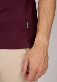 T-shirt bordeaux en coton doux, avec une fente sur le côté et une étiquette de marque à l'ourlet. Main reposant sur un pantalon beige.