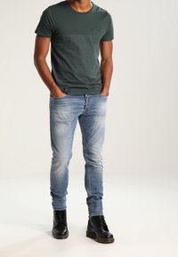 Tmavě zelené tričko s krátkými rukávy a kapsou, kombinované se světle modrými džíny a černými šněrovacími botami, stojící na neutrálním pozadí.