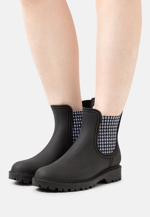 Bottes en caoutchouc noires montant jusqu'à la cheville avec des semelles épaisses à crampons et des panneaux latéraux élastiques en pied-de-poule noir et blanc, portées sur des jambes nues.