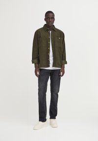 Green corduroy shirt jacket with button closure and two chest pockets, worn over a white T-shirt; paired with dark jeans and white sneakers.