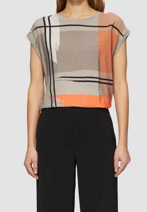 Kobieta w bluzce z krótkim rękawem z abstrakcyjnymi beżowymi, szarymi, pomarańczowymi i czarnymi blokami, zestawiona z czarnymi spodniami z wysokim stanem na szarym tle.