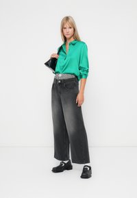Camisa verde de cetim com botão e um corte descontraído, combinada com calças de ganga largas cinzentas de cintura alta e sapatos pretos; segurando uma mala preta.