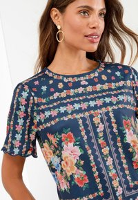 Haut à manches courtes à motif floral en bleu marine avec des manches festonnées, mettant en avant des fleurs vibrantes dans des teintes de rose, rouge et vert.