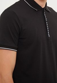 Polo noir avec un col zippé, présentant des accents blancs sur le col et les manches. Comprend la marque "Armani Exchange" le long de la fermeture éclair.