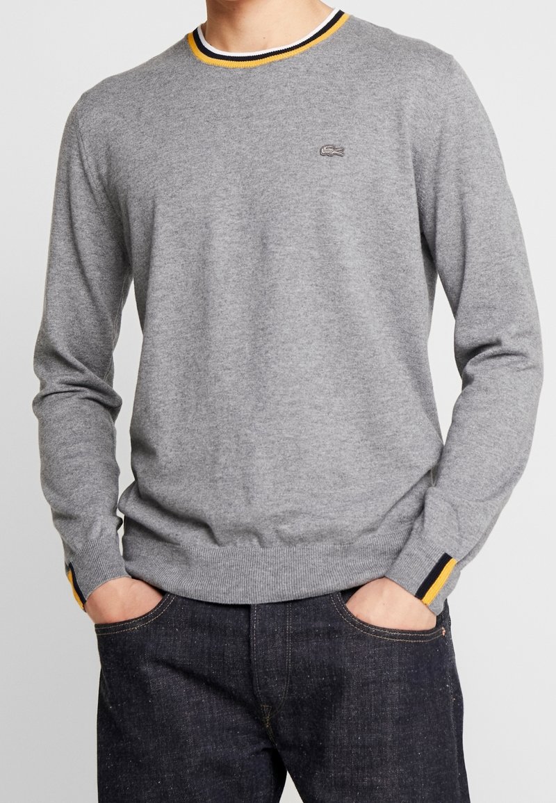 Homme portant un pull gris à manches longues avec des rayures jaunes et noires sur le col et les poignets, mains dans les poches, jean foncé.