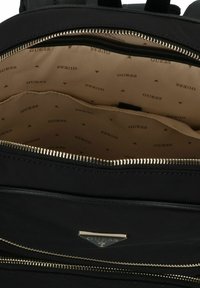 Interno di uno zaino nero in nylon con fodera beige, caratterizzato da un logo "GUESS" ripetuto. Dettagli in oro della cerniera e molteplici tasche visibili.