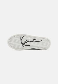 Valge sneakersole, millel on kalasabamuster, sile pind ja silmapaistev must logo keskel.