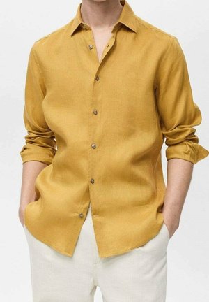 Mężczyzna w musztardowożółtej koszuli z długim rękawem, z podwiniętymi rękawami, ze złożonymi rękami i jasnobeżowymi spodenkami na jednolitym tle.