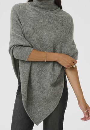 Kobieta nosząca teksturowane szare dzianinowe ponczo z golfem na długim rękawie i czarne spodnie, poprawiająca ponczo na ramieniu.