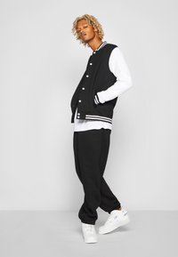 Chaqueta varsity en blanco y negro con botones a presión, puños a rayas, combinada con pantalones de chándal negros y zapatillas blancas. Textura de tela suave.