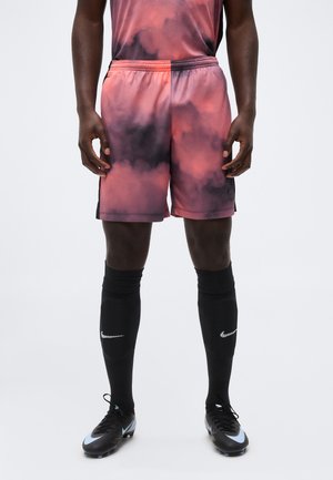 Athlète masculin portant un short tie-dye rouge et noir, des chaussettes Nike noires montantes jusqu'aux genoux et des crampons noirs sur un fond clair.