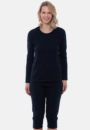 Vrouw met schouderlang blond haar die lacht, gekleed in een aangesloten marineblauw shirt met lange mouwen en marineblauwe cropped broek, staand.