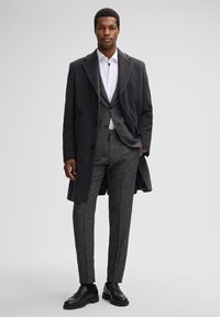 Grijze wollen overjas over een licht overhemd en een gestreept grijs pak. De outfit omvat taps toelopende broek en zwarte instapschoenen.