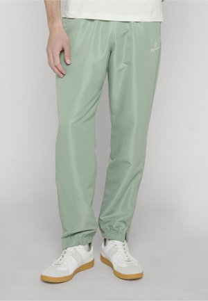 Persona che indossa pantaloni da jogging verde chiaro con polsini elastici e sneakers bianche con suole in gomma, in piedi contro uno sfondo neutro.
