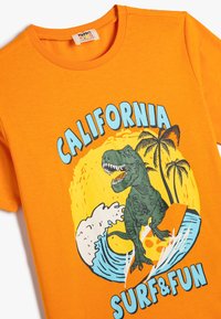 Oranssi puuvillapaita, jossa on dinosauruksen kuva surffaamassa, ja teksti "CALIFORNIA SURF & FUN" sekä palmupuuyksityiskohdat sinisellä ja keltaisella.