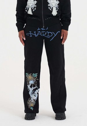 Pantalons de survêtement noirs avec le texte bleu "ED HARDY", présentant une broderie complexe de dragon et de fleurs en bleu, vert et jaune sur la jambe gauche.