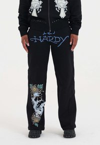 Zwarte joggingbroek met blauwe "ED HARDY" tekst, voorzien van ingewikkelde draak- en bloemenborduursels in blauw, groen en geel op het linkerbeen.