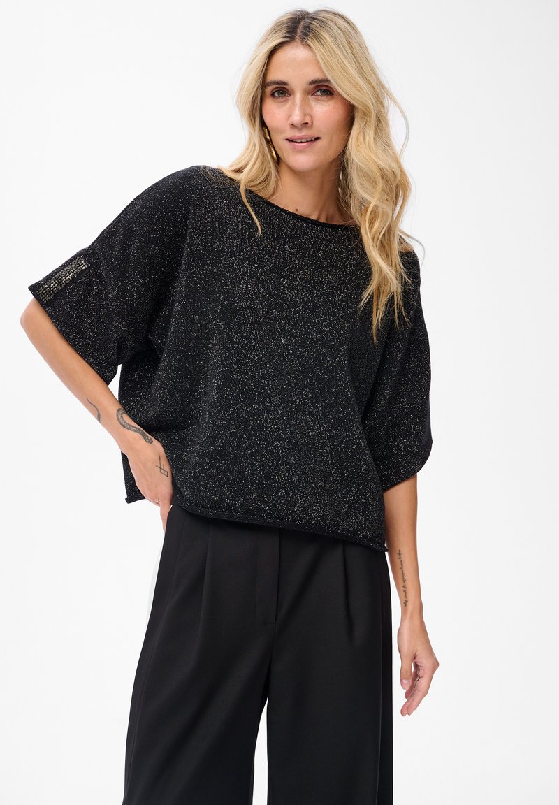 Czarny oversize sweter z metalicznymi akcentami, szerokim dekoltem w łódkę, krótkimi rękawami oraz wyraźnie texturedowanym materiałem. Zestawiony z czarnymi spodniami.