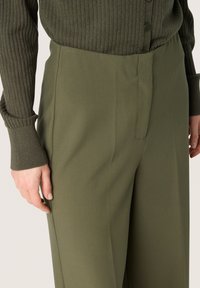 Olivgrüne, hochtaillierte Hose mit glatter Textur und Seitentasche. Kombiniert mit einem strukturierten, passenden Langarmoberteil, ebenfalls olivgrün.