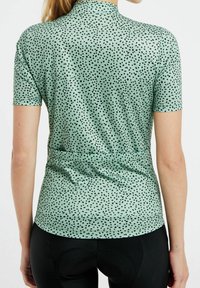 Chemise verte à manches courtes avec un motif moucheté noir, un col haut et une coupe ajustée avec un détail de lien à la taille.