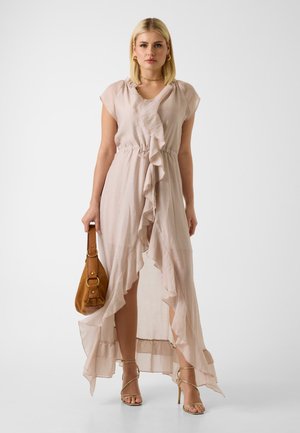 Donna con un vestito rosa chiaro a volant, che tiene una borsa marrone, indossa sandali dorati con cinturini, in piedi su sfondo bianco.