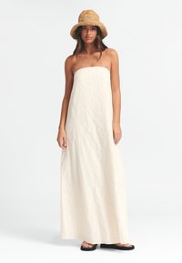 Ärmelloses, cremefarbenes Maxikleid mit vertikaler Rippstruktur. Das Modell trägt einen Strohhut und schwarze Flip-Flops und steht vor einem hellen Hintergrund.