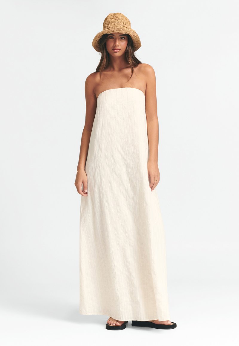 Brezramena, krema barve maksi obleka z navpično rebrasto teksturo. Model nosi slamnati klobuk in črne japonke, stoji ob svetli ozadju.