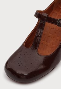Chaussure à bride en T en cuir verni marron foncé avec de petites perforations sur l'avant et une semelle intérieure rembourrée, vue en plongée inclinée.