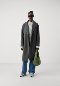 Szürke gyapjú kabát, világosszürke kötött pulóver, kék farmernadrág és ezüst sportcipők. Texturált zöld válltáskát tart, napszemüveget visel.