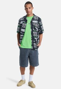 Πουκάμισο με κοντά μανίκια σε μαύρο χρώμα με λευκά και πράσινα γραφικά μοτίβα, σε συνδυασμό με φωτεινό πράσινο μπλουζάκι με στρογγυλή λαιμόκοψη και τζιν σορτς.