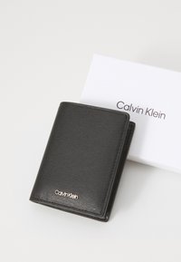 Musta nahkalompakko, jossa on tekstuuri ja kultainen "Calvin Klein" -logo. Mukana valkoinen laatikko, jossa on painettu brändin nimi.