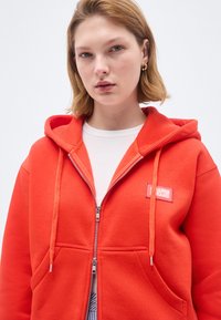 Tânără cu păr blond scurt poartă un hanorac portocaliu aprins cu fermoar peste un top alb, privind drept înainte cu o expresie neutră.