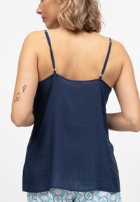 Kobieta z krótkimi blond włosami, ubrana w granatową bluzkę bez rękawów i wzorzyste niebiesko-białe spodnie, pokazująca swoje tatuaże na plecach i ramionach.