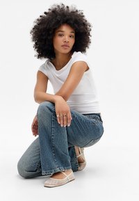 Ung kvinde med krøllet hår, der knæler, iført en hvid tætsiddende t-shirt, blå stribede jeans, ringe og beige net-sandaler mod en hvid baggrund.