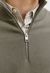 Pull en maille vert olive avec un col zippé, doté d'une fermeture éclair argentée et d'une chemise blanche rayée en dessous. Design texturé et décontracté.