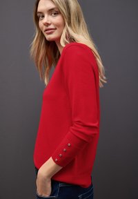Roter Strickpullover mit langen Ärmeln, der Knopfakzente an den Bündchen aufweist. Glatte Textur und figurbetontes Design, kombiniert mit blauen Jeans.