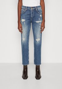 Jeans de mezclilla azul con detalles desgastados, diseño de corte recto, arrugas y parches descoloridos. Llevados con botas de tobillo marrones.