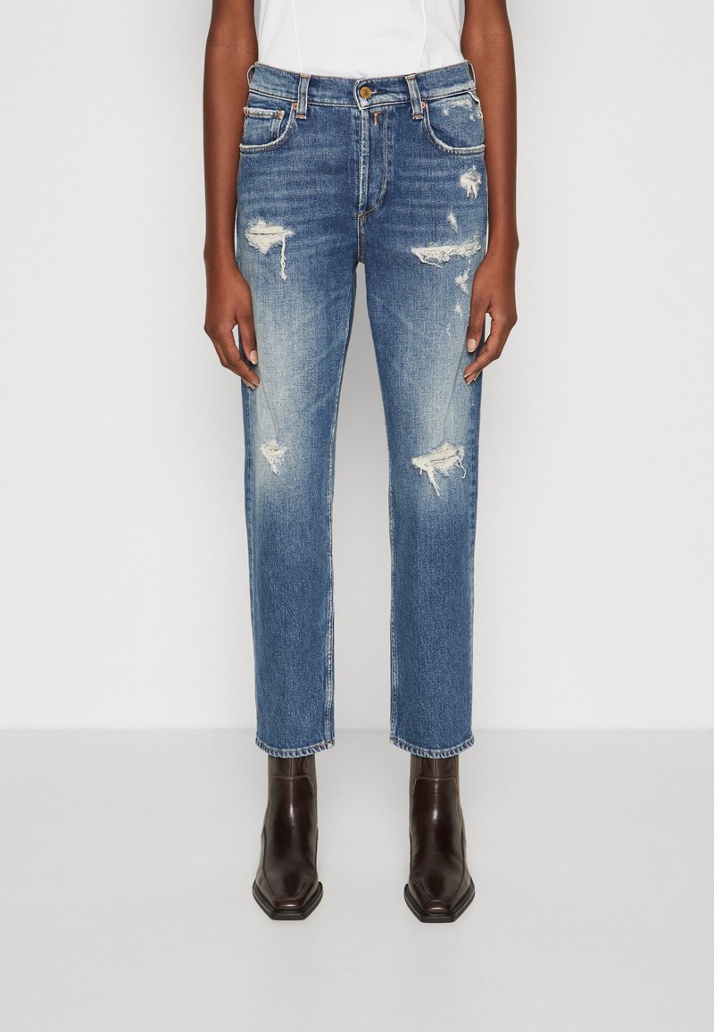Jeans de mezclilla azul con detalles desgastados, diseño de corte recto, arrugas y parches descoloridos. Llevados con botas de tobillo marrones.