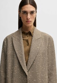 Houndstooth-patterned blazer in beige and brown with a textured finish, paired with a collared brown shirt. Features wide lapels and a tailored fit.