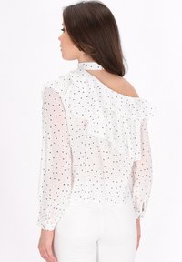 Weiße Bluse mit schwarzen Punkten, Rüschendesign entlang der Schultern, durchsichtige lange Ärmel und mit Knöpfen an den Bündchen. Offener Rücken mit einer Schleife am Hals.