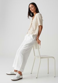 Beige short-sleeve cardigan with a textured pattern, featuring a V-neckline and front ties, paired with loose white trousers and white sneakers.