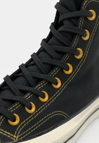 Zapatillas de lienzo de caña alta negras con ojales amarillos, cordones negros y costuras amarillas; suela de goma con un acabado texturizado.