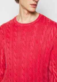 Maglione rosso a maglia con collo rotondo, caratterizzato da modelli in rilievo e texture a maglia. Il tessuto appare morbido e leggero.