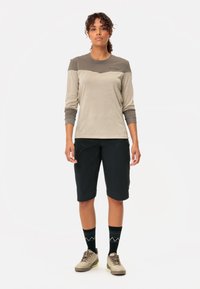 Chemise à manches longues de couleur beige clair, évacuant l'humidité, avec des accents gris foncé, associée à un short noir et des chaussettes noires à motifs, le tout complété par des chaussures beiges.