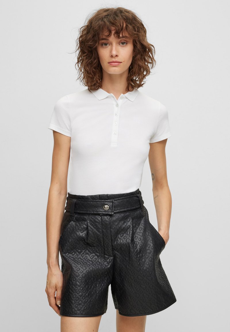 Biała koszulka polo z kołnierzykiem i zapięciem na guziki, zestawiona z wysokim stanem, teksturowanymi czarnymi szortami o wzorze przypominającym skórę.