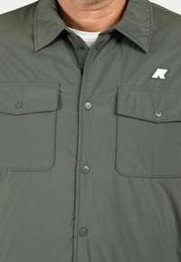Camicia grigia con bottoni, realizzata in tessuto leggero; presenta due tasche sul petto, bottoni a pressione verdi e un piccolo logo sul lato sinistro.