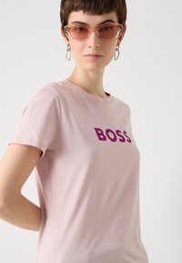 Šviesiai rožinė T-palaidinė su ryškiu violetiniu "BOSS" logotipu priekyje, trumpomis rankovėmis ir apvaliu iškirpimu, pagaminta iš švelnaus medvilnės audinio.
