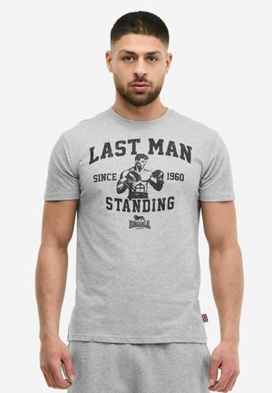 Mann mit kurzem dunklem Haar und Bart, trägt ein graues T-Shirt mit schwarzem Boxmotiv und dem Text "Last Man Standing Since 1960 Lonsdale London".